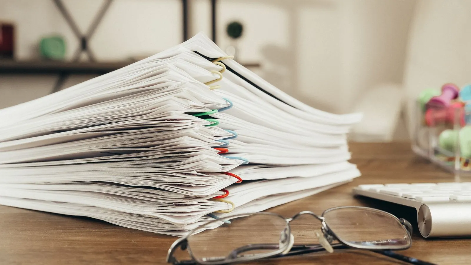 A tall stack of paper-clipped documents on a wooden desk beside reading glasses and a keyboard