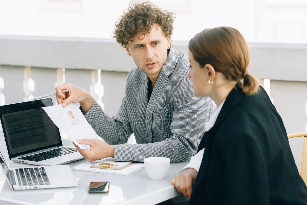 Two colleagues at an outdoor table with laptops, one holding up a printed chart while explaining it