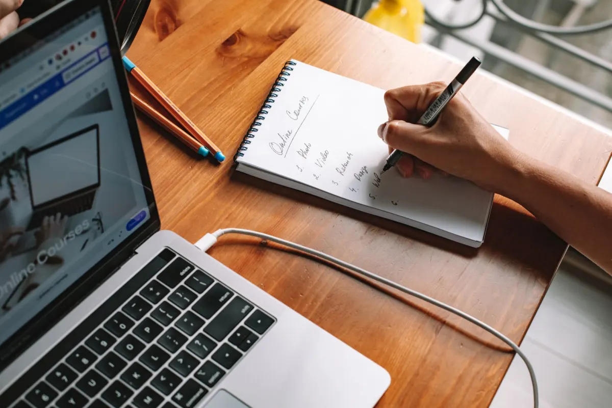 A hand writing a numbered list headed Online Courses in a spiral notepad beside an open laptop