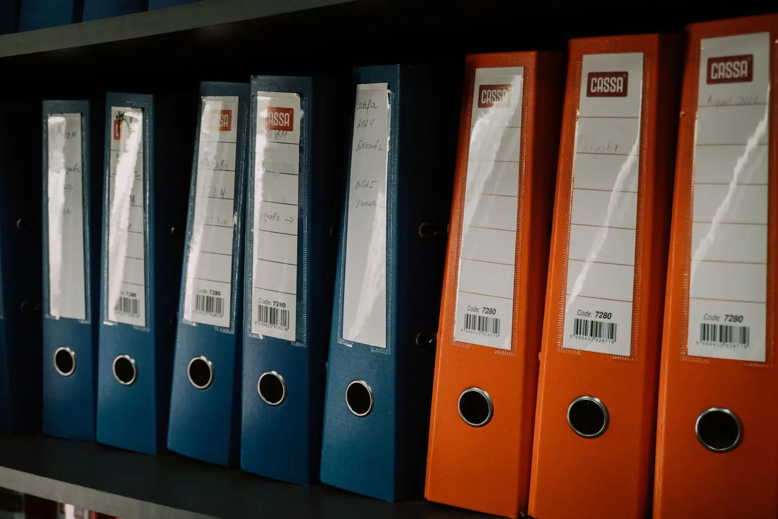 Rows of blue and orange ring binders with handwritten labels on an office shelf