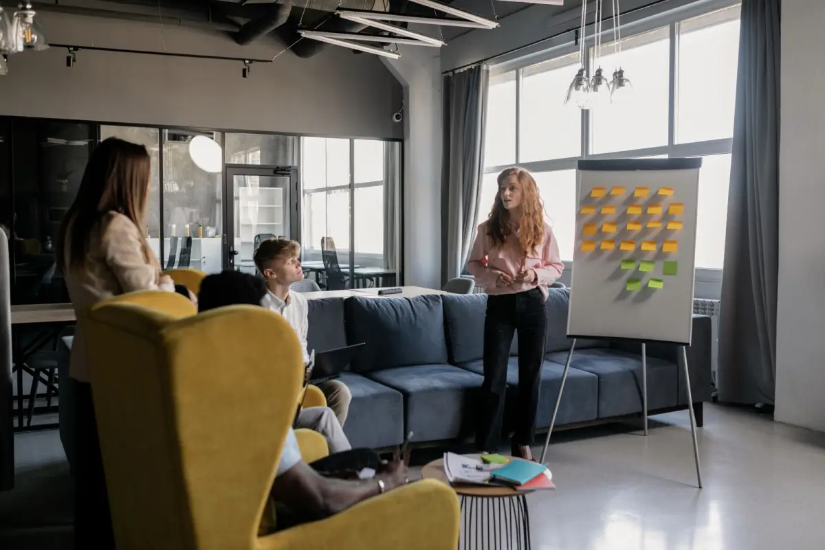 A woman stands beside a flip chart of colored sticky notes while three colleagues listen from armchairs and a sofa